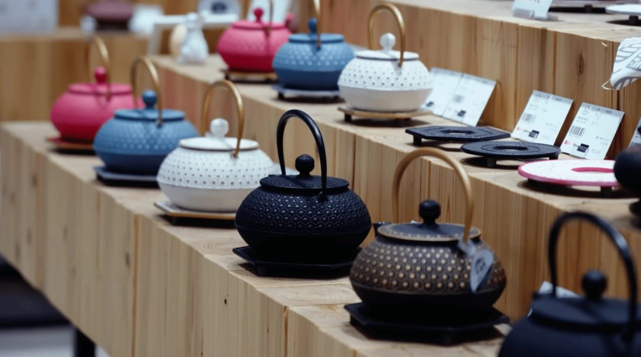Rows of small cast iron teapots on stepped wooden shelves in pink, blue, white, black and bronze, each with an arched handle and studded surface.