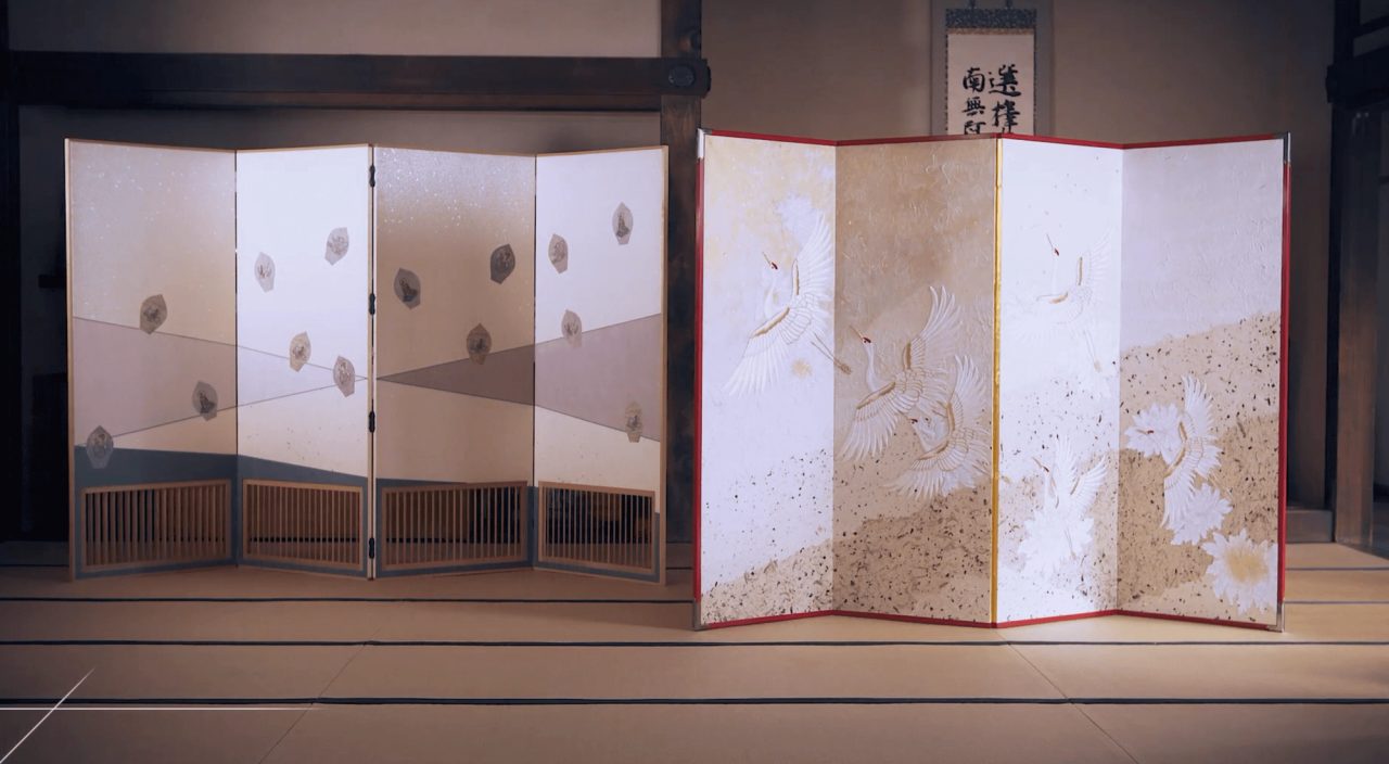 Two folding screens on a tatami floor, one pale with scattered fan shapes, one with white cranes on a gold ground within a red frame.