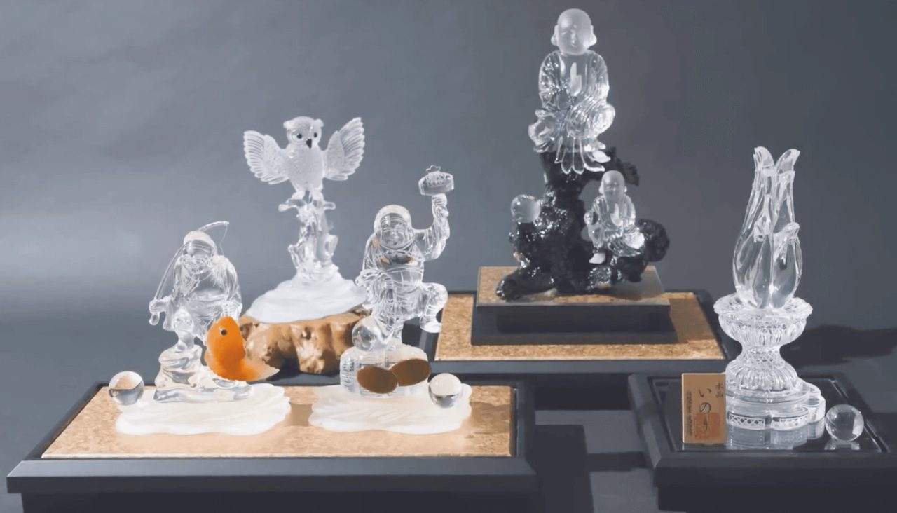 Carved clear crystal figures on stone plinths against a grey background, including an owl, seated figures, and a lotus-shaped vessel.