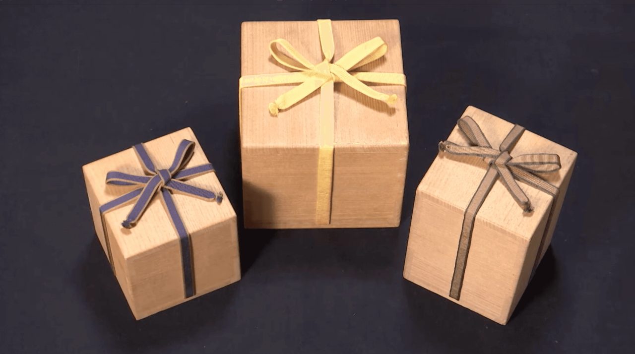 Three plain wooden boxes on a dark surface, each tied with a flat woven tape in cream, indigo or grey, finished in a flat bow.