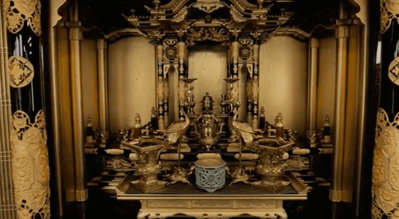 The interior of a household Buddhist altar in gold lacquer, its tiered miniature hall filled with columns, carved eaves and metal offering vessels.