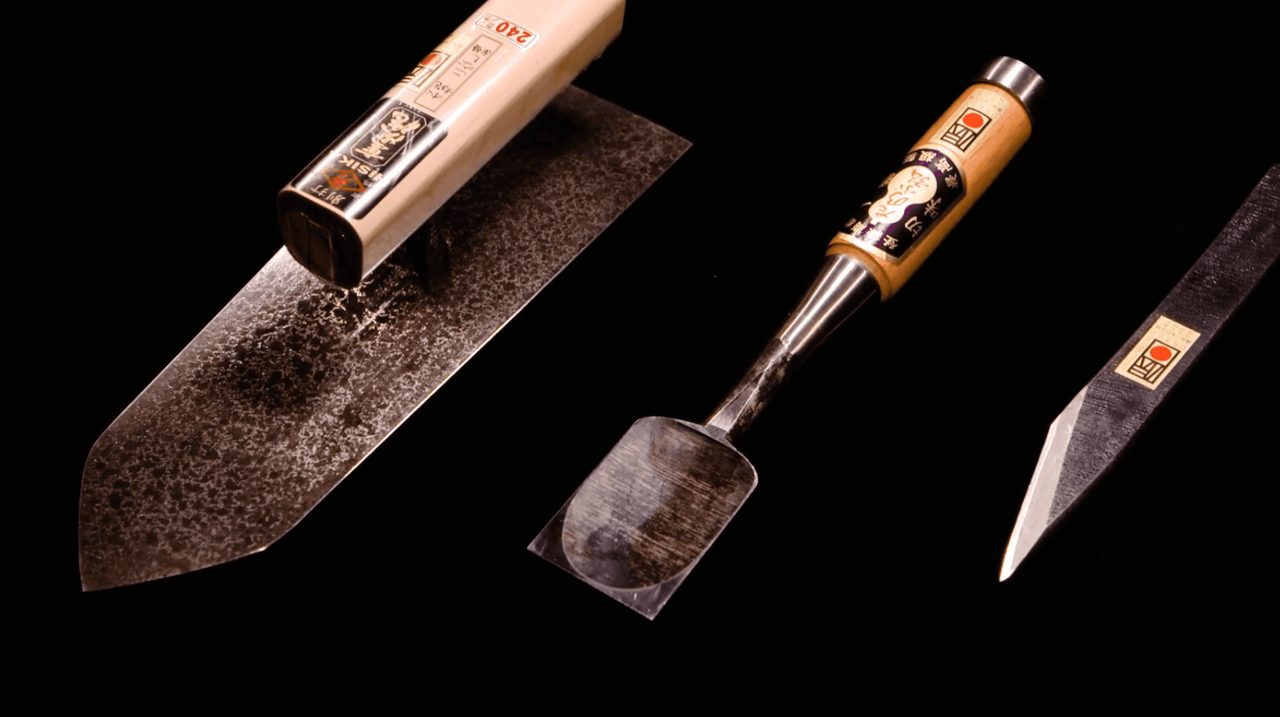 Three forged tools on a black background, a broad knife with a hammered surface, a chisel with a pale wooden handle, and a narrow blade.