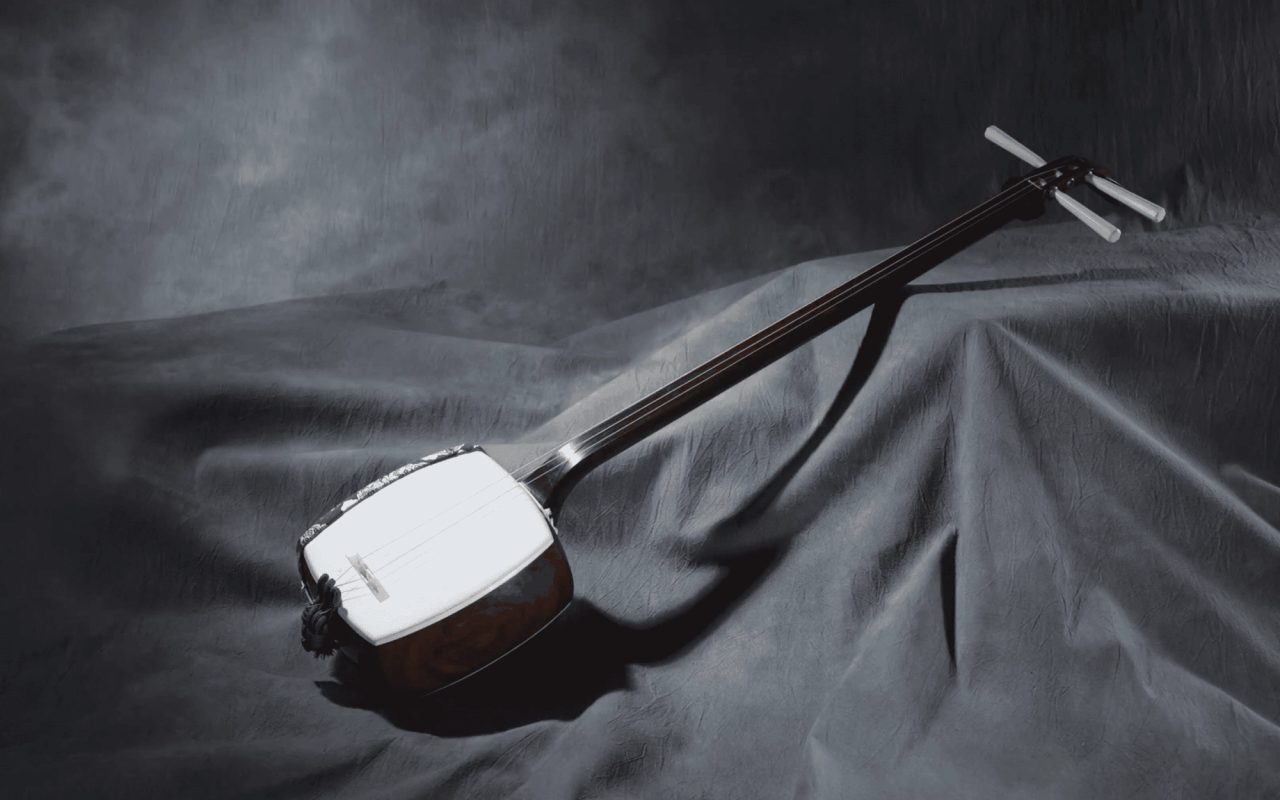 A three-stringed shamisen laid on grey draped cloth in a dark studio, its white skin-covered body and long slender neck lit from above.