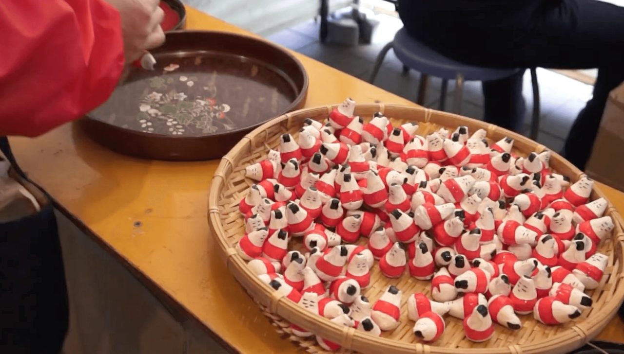 A wide flat basket heaped with small red and white roly-poly dolls, beside a lacquer tray where someone is painting faces onto more of them.