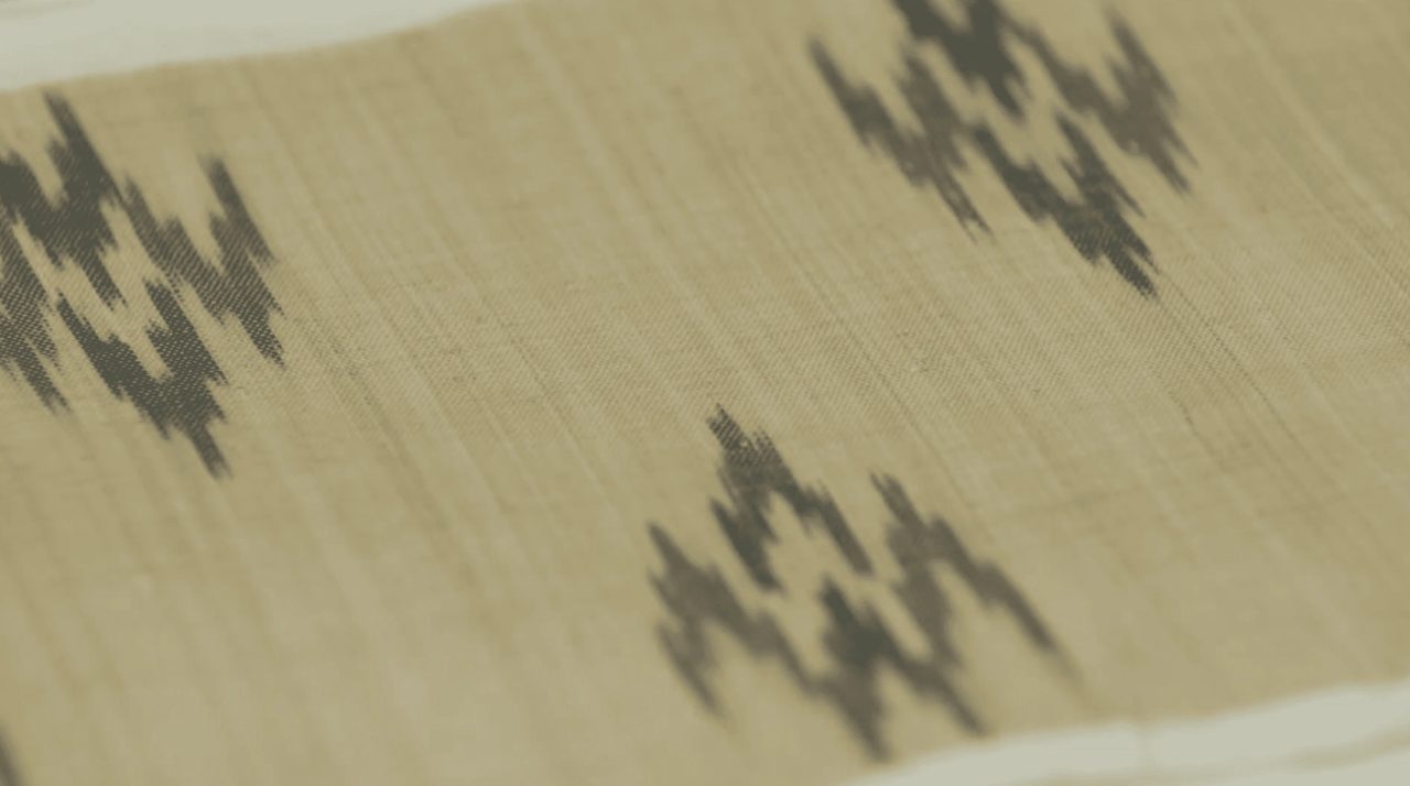 A close view of pale banana-fiber cloth, woven with dark stepped ikat motifs at intervals across the loose open weave.