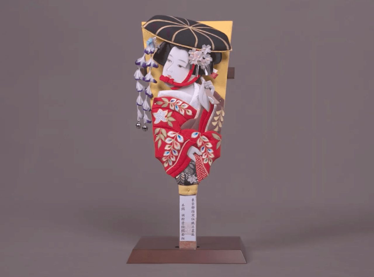 A decorated wooden paddle standing on a metal base, its face built up in padded silk as a figure in a red kimono under a black umbrella.