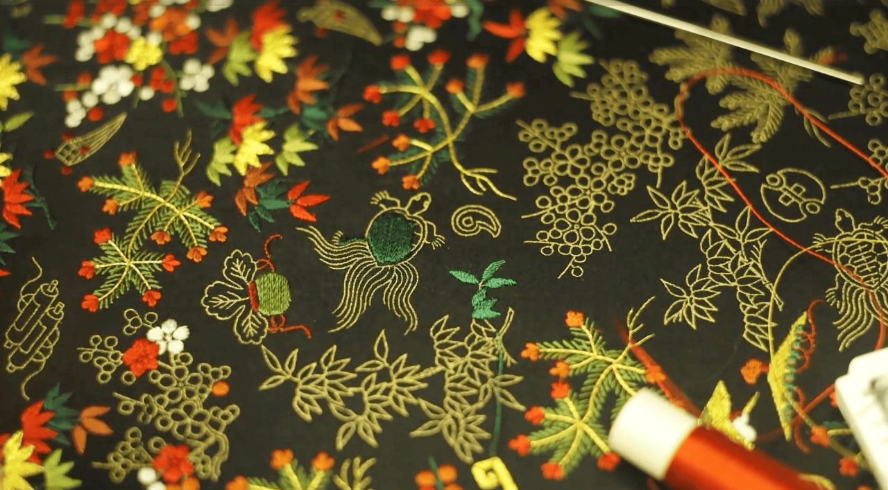 Black silk embroidered in gold, green, red and white with grasses, gourds and flowers, a needle and red thread visible at the right edge.