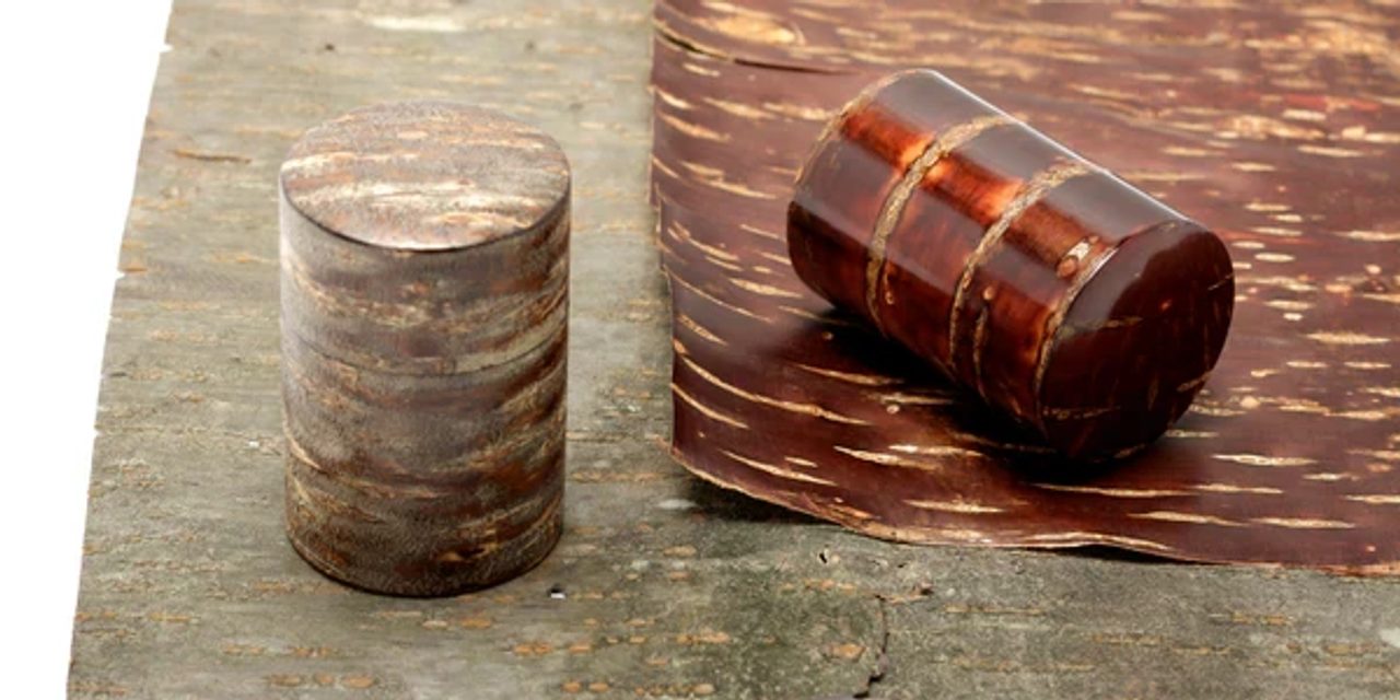 Two cylindrical tea caddies covered in polished cherry bark, one matte and pale, one glossy and dark red, on a weathered wooden plank.
