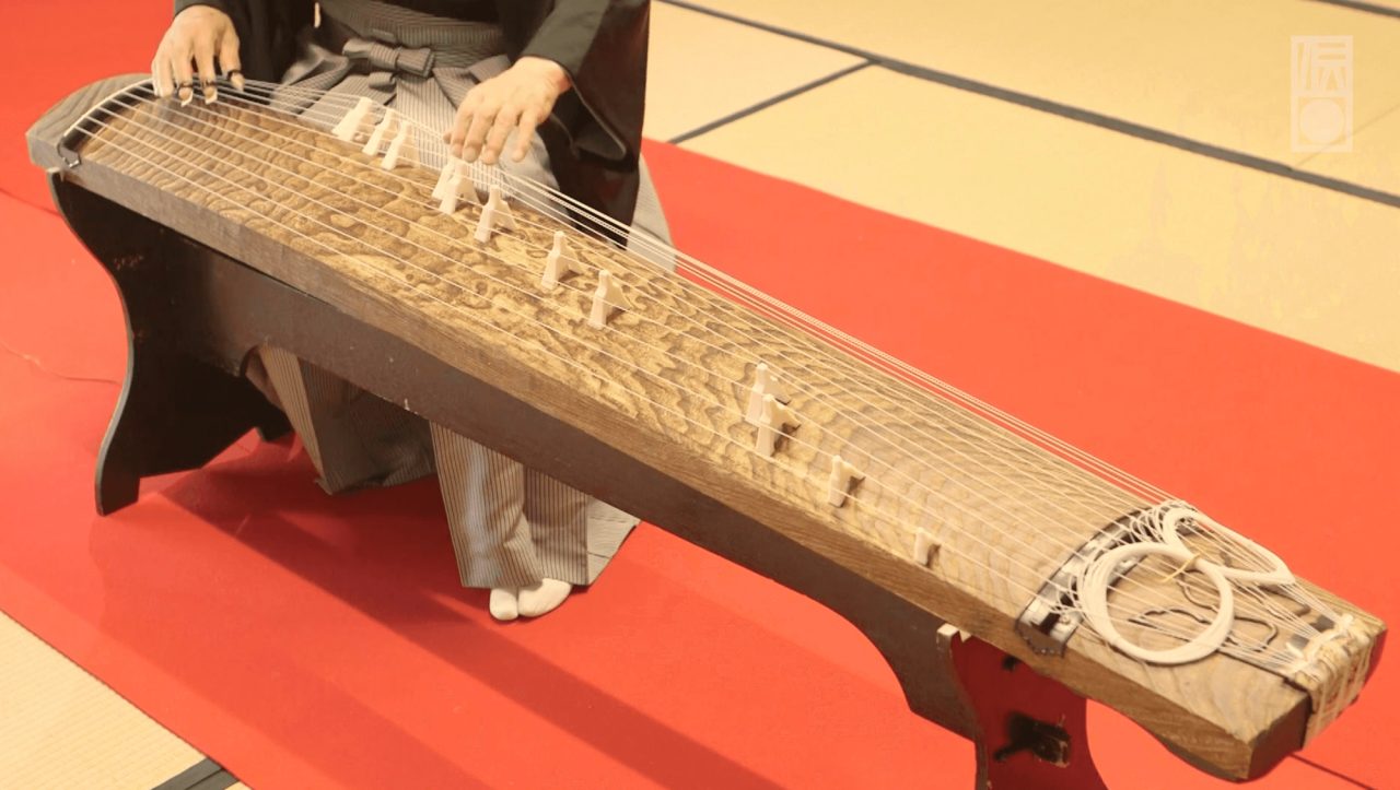 A person in dark robes seated at a long wooden koto on a red mat, fingers on the strings, the movable bridges standing in a line along the body.