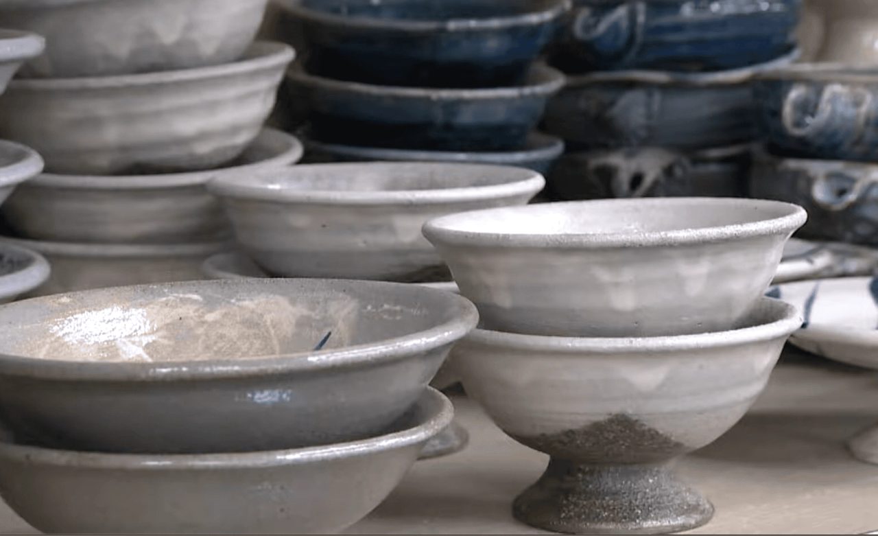 Stacks of glazed bowls and footed cups on a shelf, most in a mottled grey-white glaze, with dark blue pieces stacked behind them.