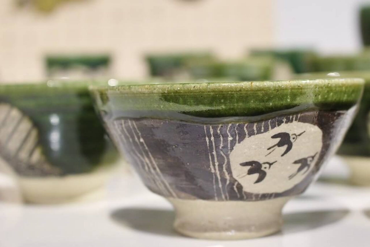 A tea bowl with a green glazed rim over a dark purple body, painted with three white birds in a pale roundel, more bowls behind it.