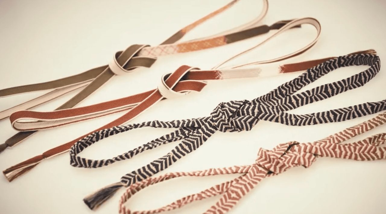 Several flat braided silk cords laid across a pale surface, some plain in olive and rust, others in black or pink chevron patterns.