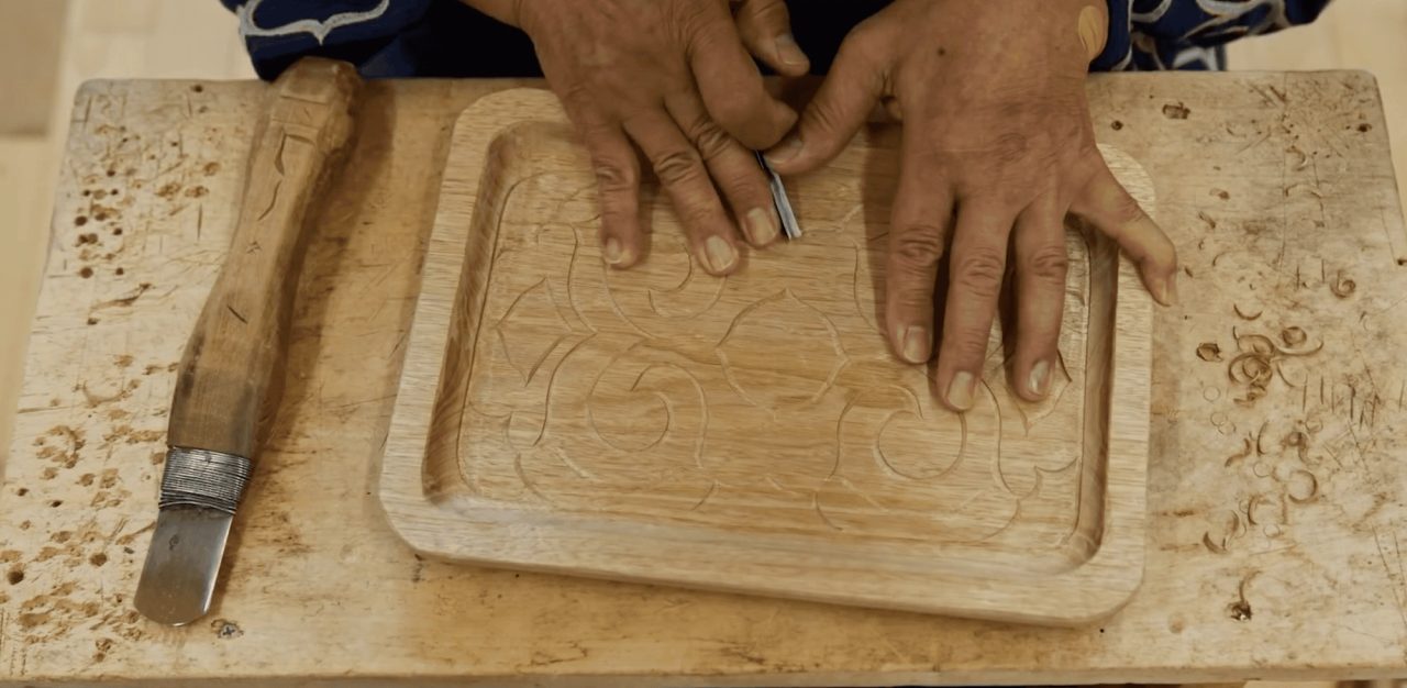 Two hands steadying a wooden tray on a workbench while a blade cuts a flowing pattern into its base, a carving knife resting alongside.