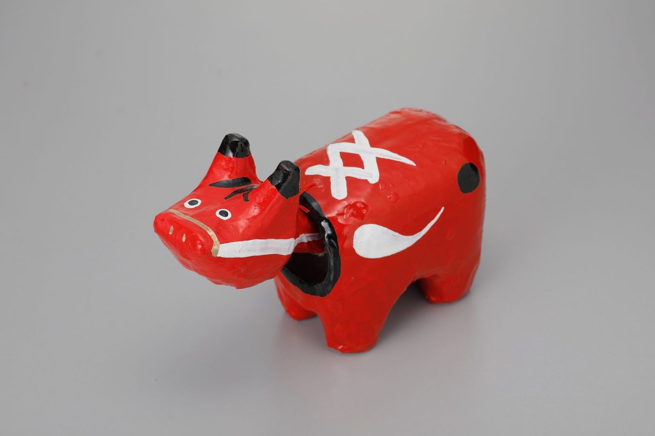 A small red papier-mache cow with a black nose and white markings, its head hanging on a separate pivot, on a plain grey background.