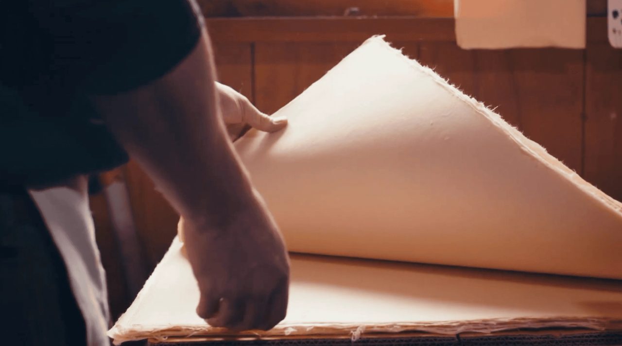 A person lifting a large damp sheet of handmade paper from a stack, the sheet curving as it comes away in a warmly lit workshop.