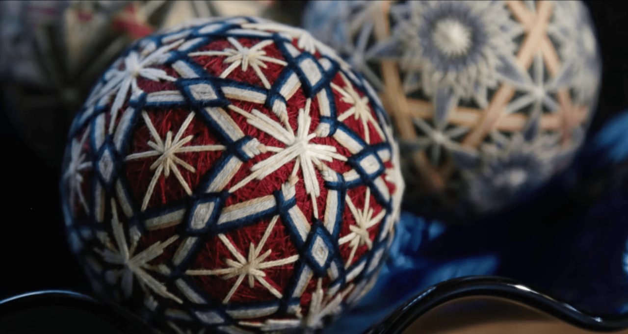 An embroidered thread ball in deep red, wrapped in white and indigo stars and diamonds, with more balls out of focus behind it.