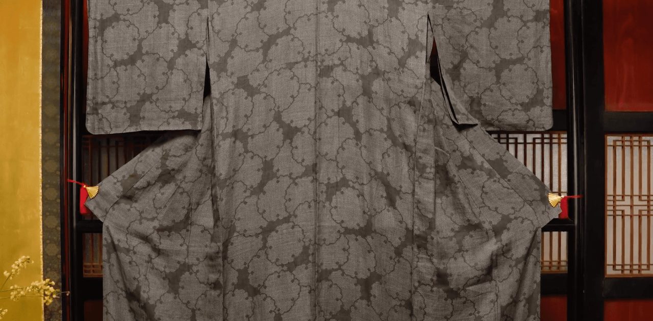 A grey kimono displayed flat on a stand, woven with a fine looping pattern, before dark wooden latticework and a gold screen.