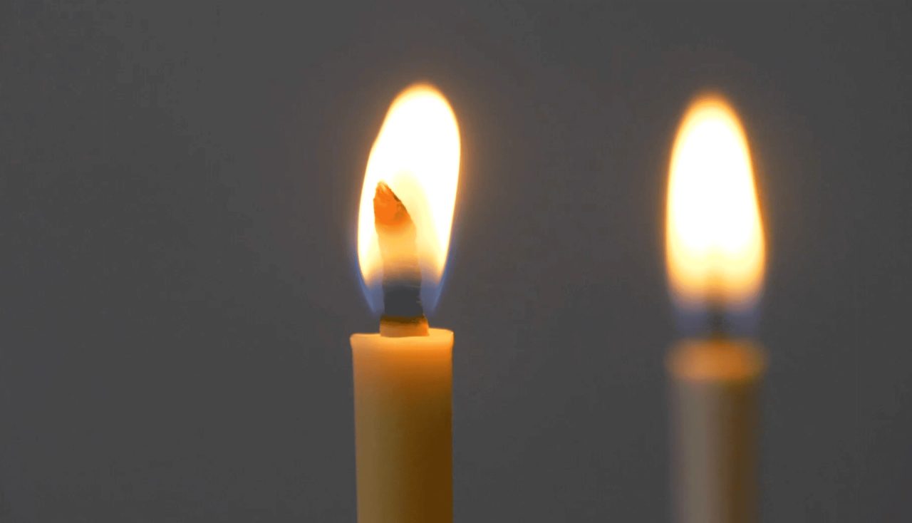 Two candles burning against a grey background, each with a tall pointed flame rising from a thick hollow wick.