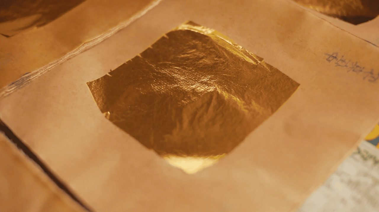 A square sheet of gold leaf lying on brown paper, its surface creased and catching the light, with more interleaved sheets around it.