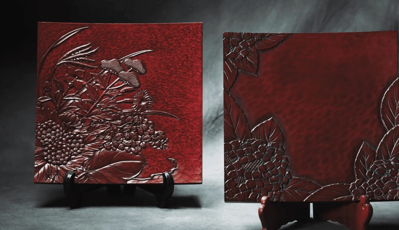 Two square carved lacquer panels on stands, deep red, one cut with chrysanthemums and grasses, the other with leaves along its edges.