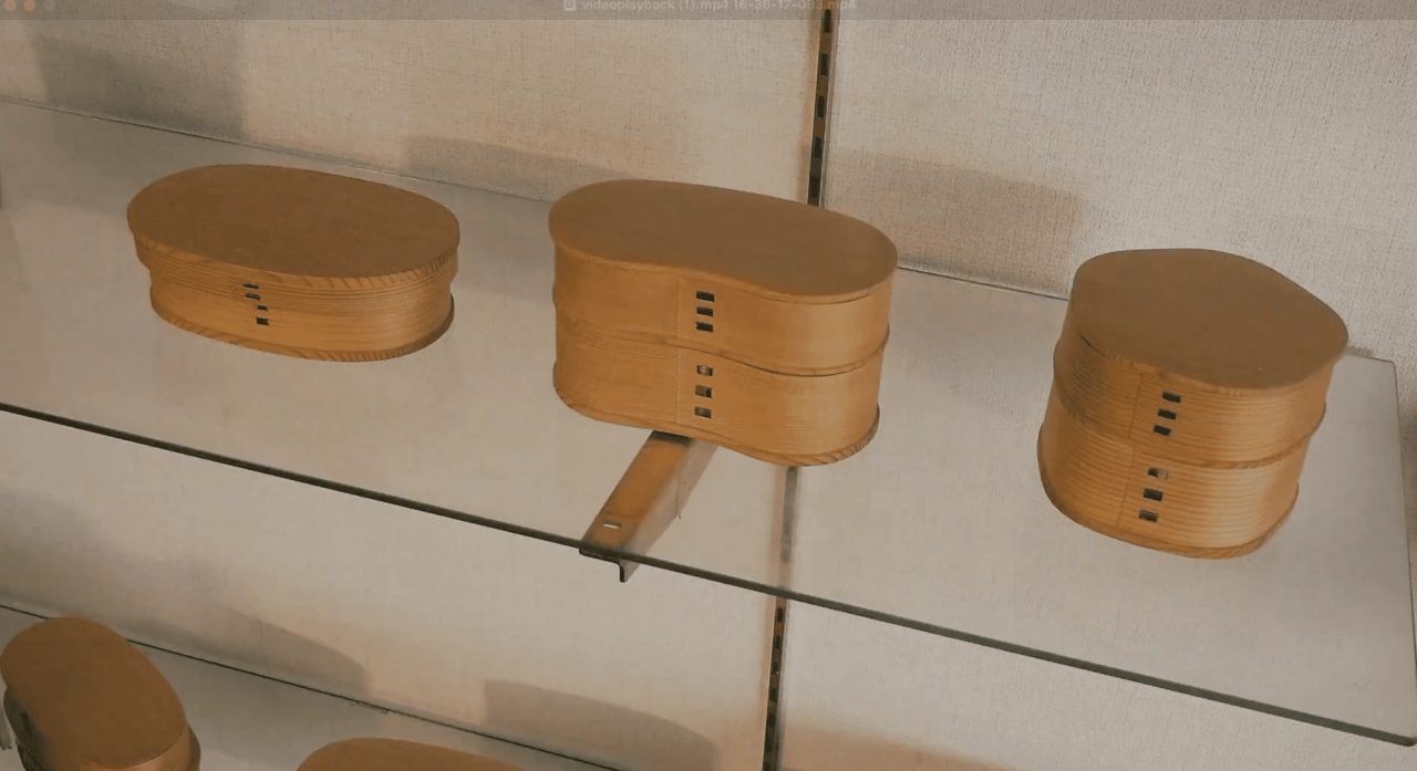 Bentwood cedar lunch boxes displayed on glass shelves, oval and gourd-shaped, each seam stitched with fine dark bark thread.