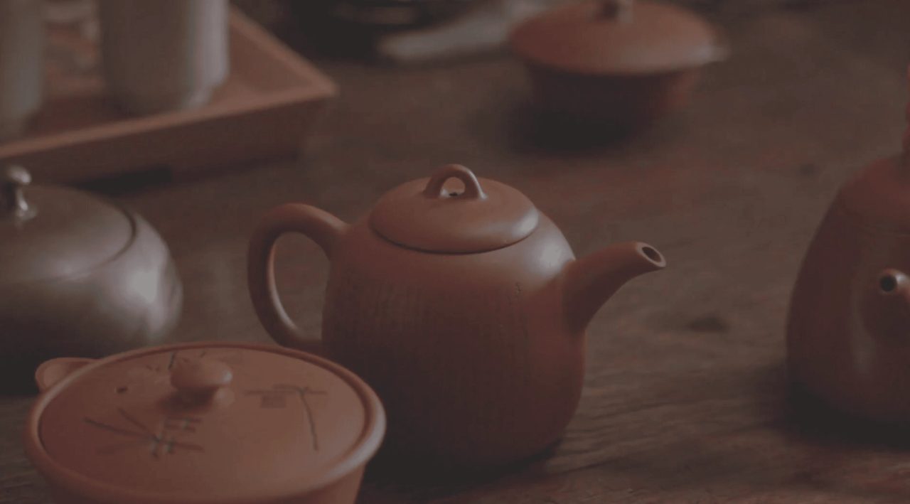 An unglazed reddish-brown clay teapot with a side handle on a wooden table, with more pots and lids out of focus around it.