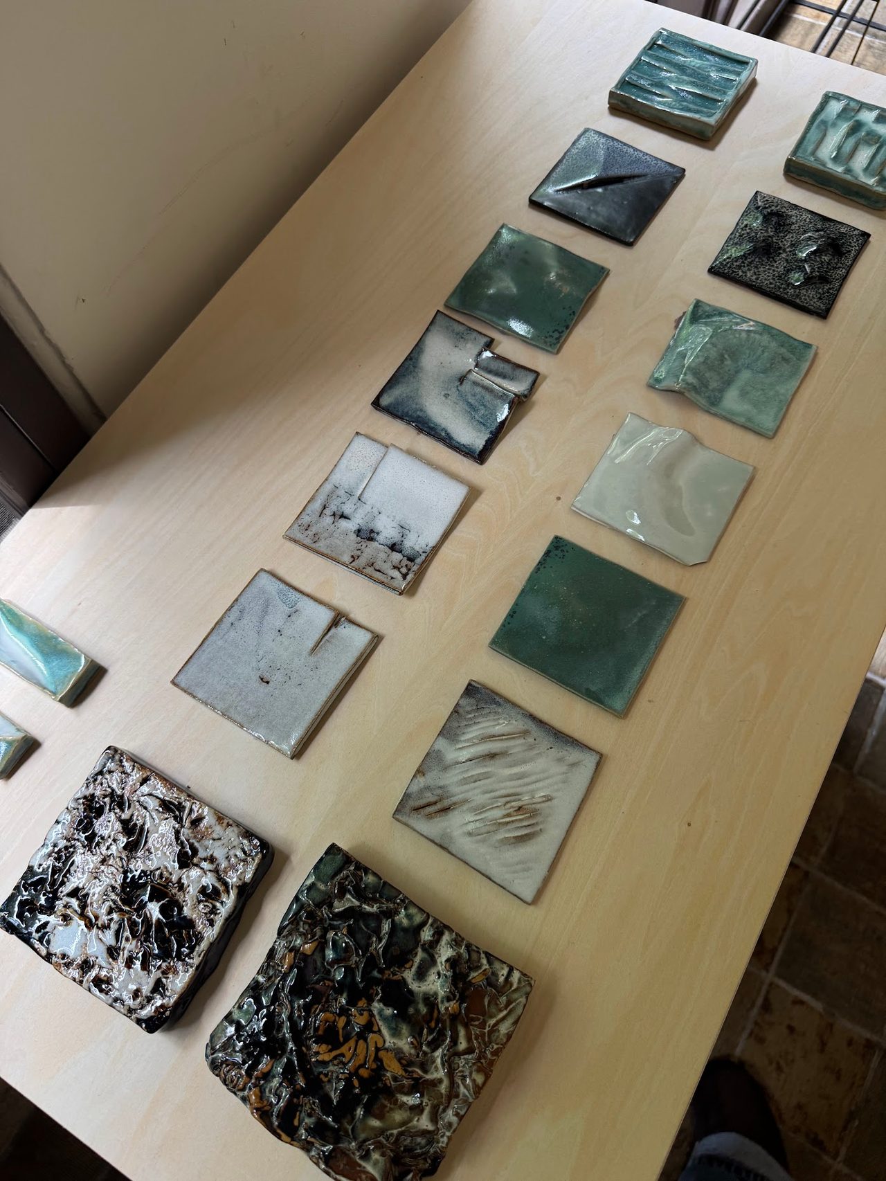 Glazed tile samples laid out on a workbench in greens, greys and speckled black.