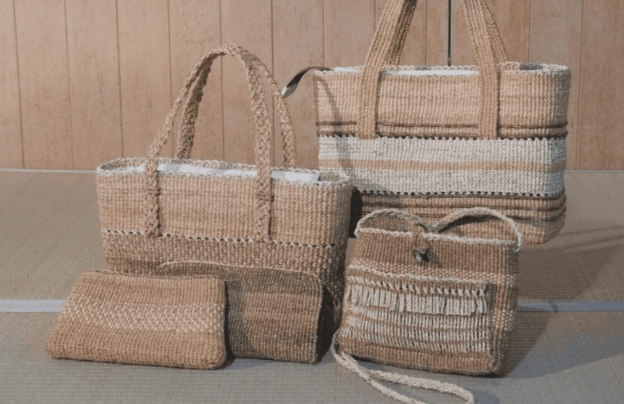 Five bags and pouches woven from pale vine and bark fiber, standing on tatami against a wooden wall, in plain and banded weaves.