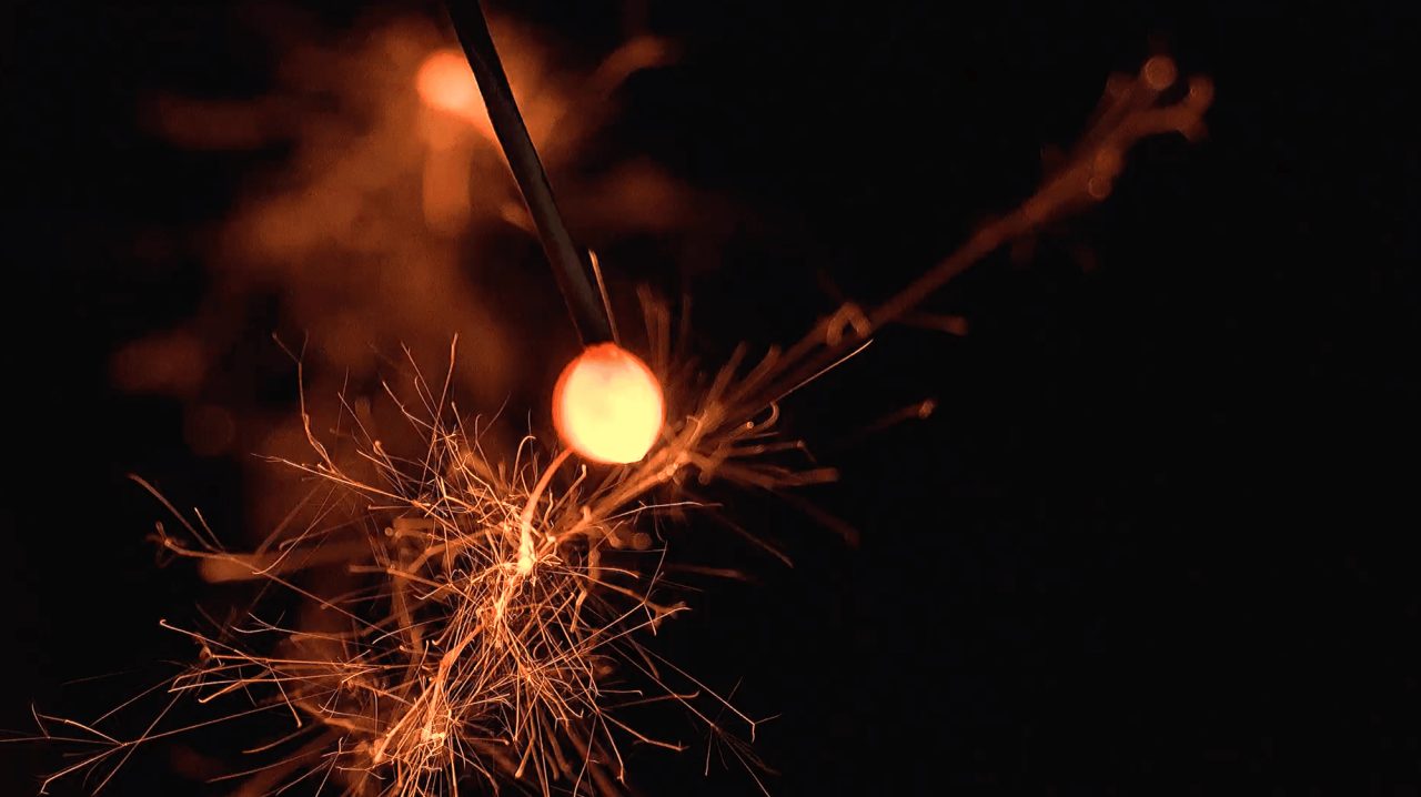 A hand-held sparkler burning at the end of a thin stick, its molten bead throwing fine orange filaments outward in the dark.