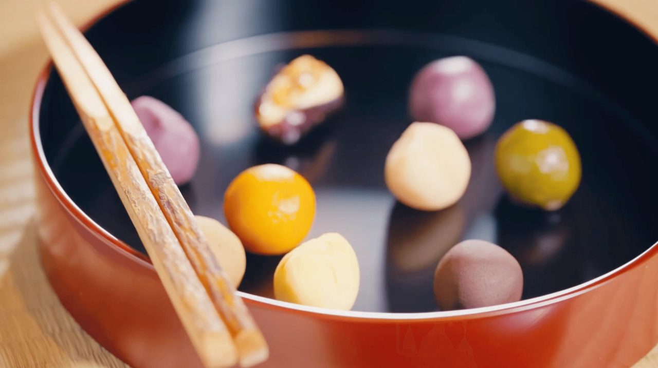 A red and black lacquer bowl holding small round sweets in purple, orange, yellow, green and brown, with a pair of wooden chopsticks resting across the rim.