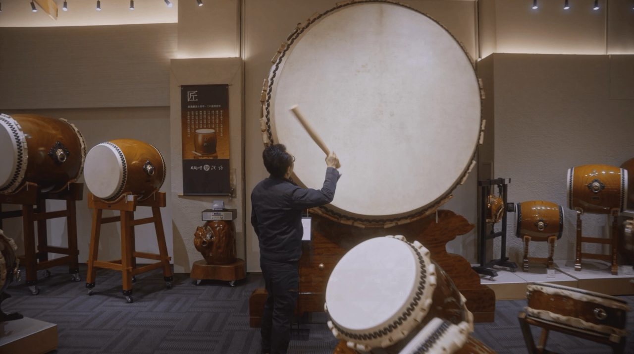 A person in a workshop showroom striking a very large drum mounted on a stand, with smaller drums on stands around the room.