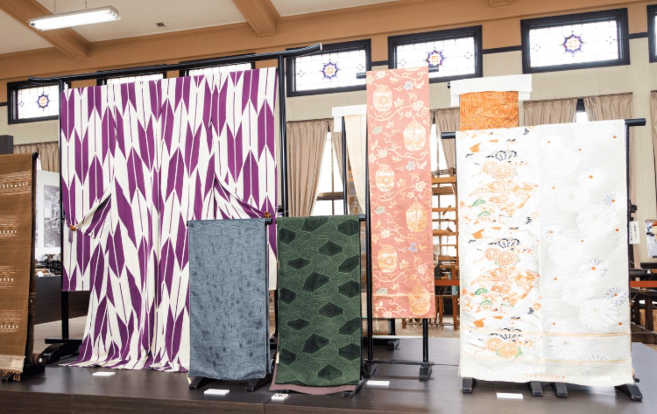 Seven lengths of woven silk displayed upright on stands in a hall, in purple and white, green, coral, and white with painted motifs.