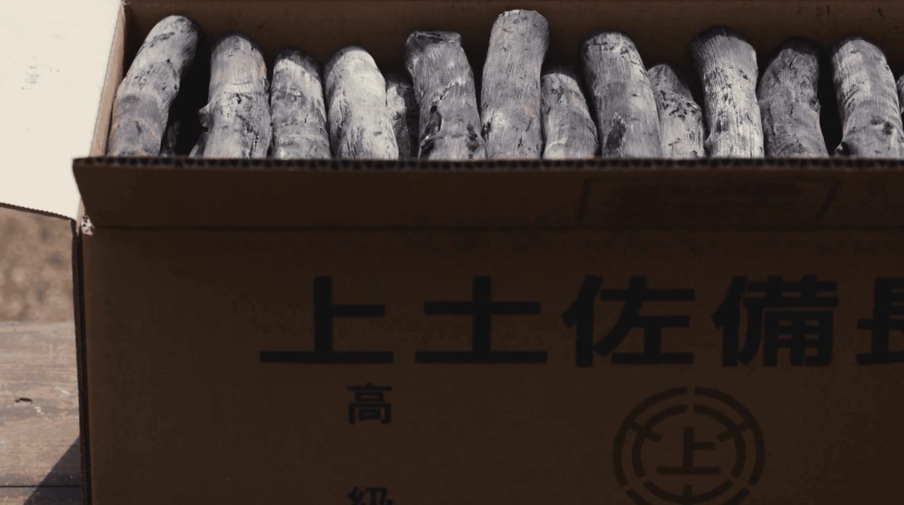 A cardboard box printed with Japanese characters, packed upright with pale grey sticks of hard charcoal.