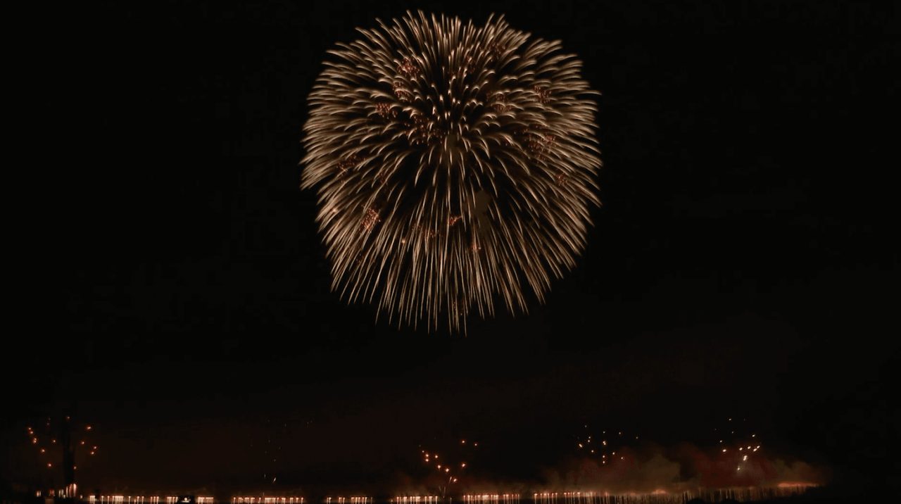 A single large firework opening as a pale gold sphere high over a dark river, with lights along the far bank reflected in the water.