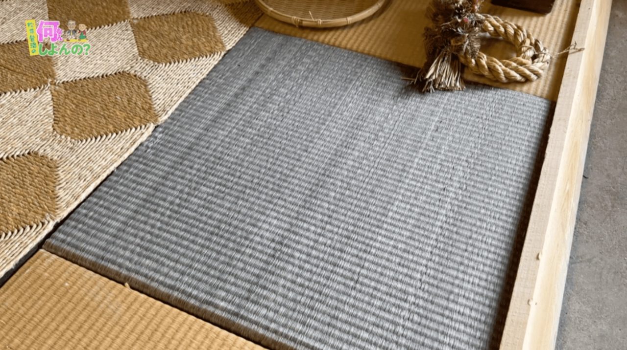 A grey woven rush mat laid inside a wooden frame on tatami, beside a checked mat in natural and brown and a coil of straw rope.