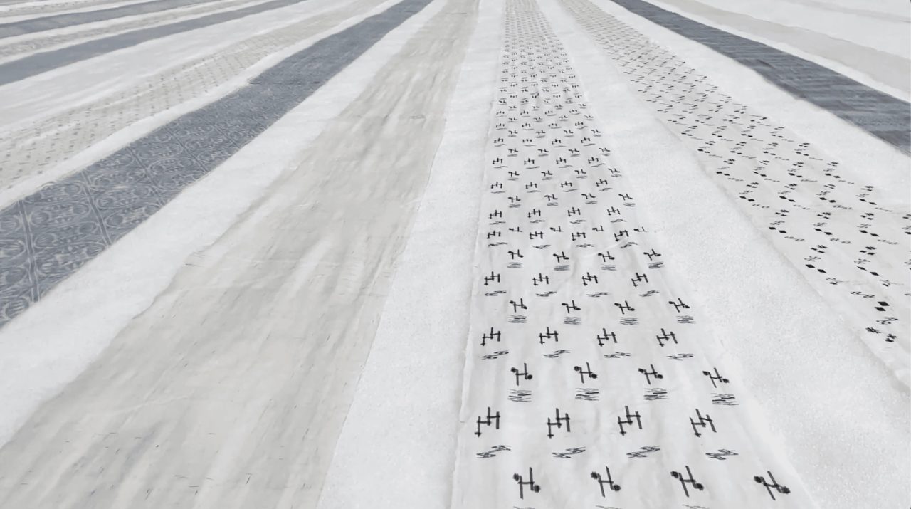 Lengths of white cloth laid flat on snow to bleach, some plain, some patterned with small dark motifs, running away from the camera in stripes.