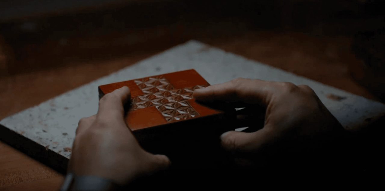 Two hands holding a small red and black lacquered box carved with a repeating geometric pattern, over a stone slab in low light.