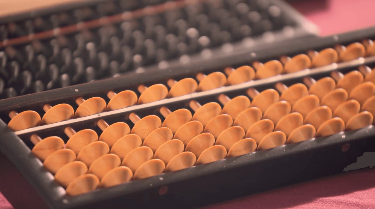 A close view of an abacus, its pale wooden beads threaded in rows on a dark frame, with another resting behind it.
