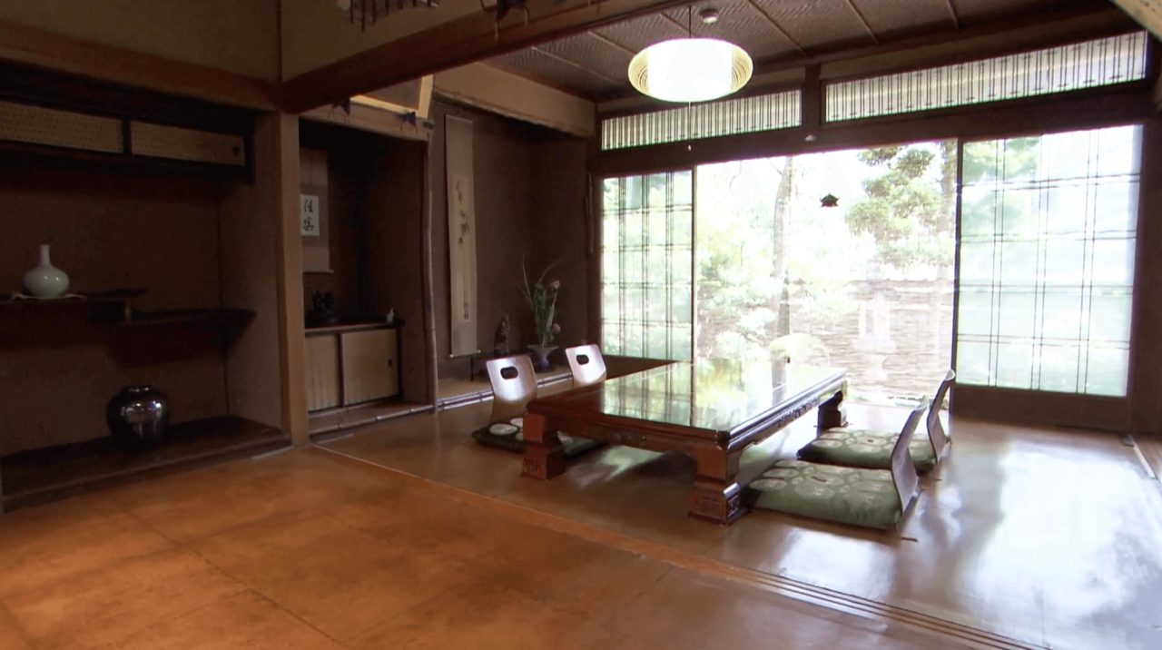 A traditional room with a low table and floor cushions, a paper lantern above, and an unbroken oiled paper floor covering running to the garden doors.