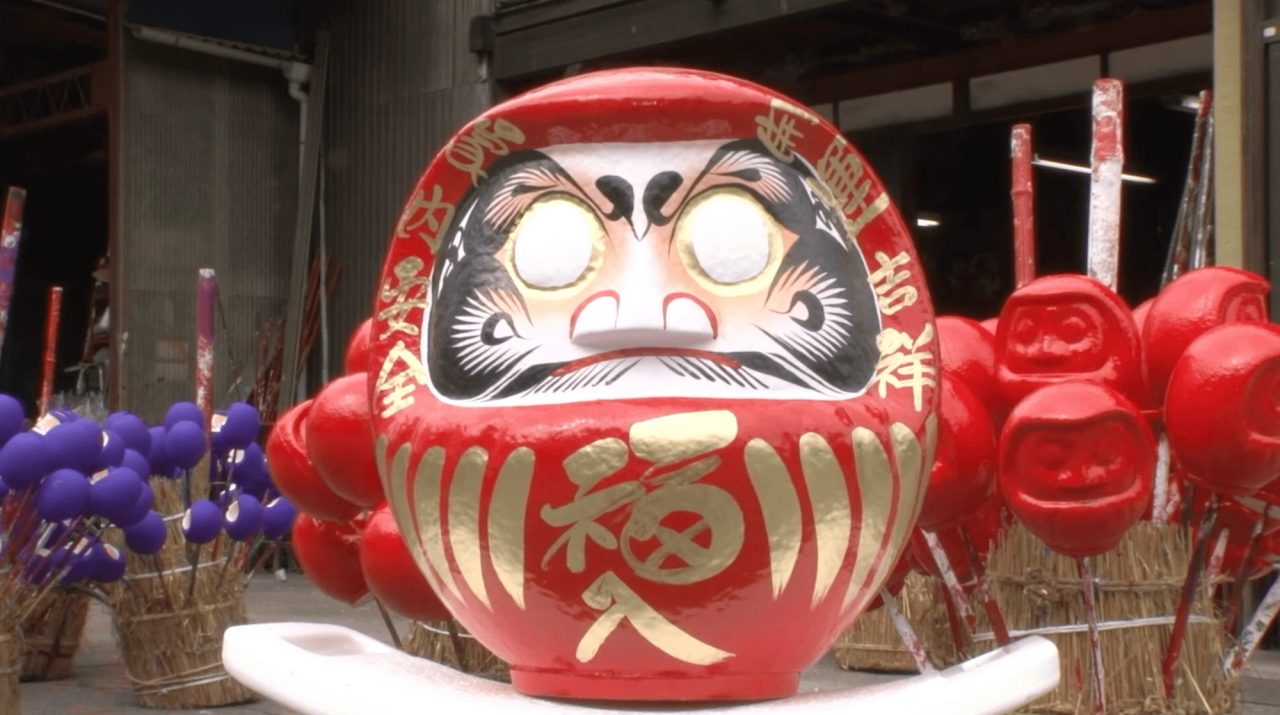 A large red daruma figure with a painted face and gold characters, standing outside a workshop among stacks of smaller red and purple ones.