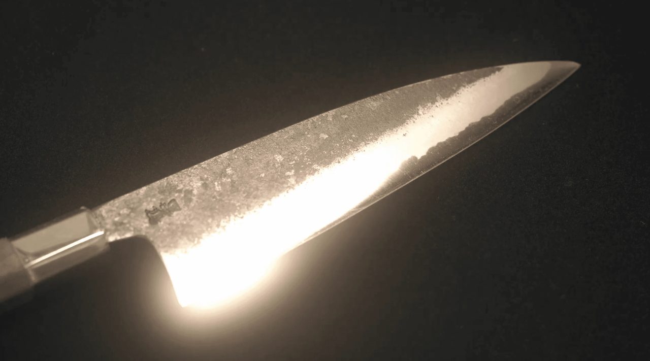 A forged kitchen knife blade lit against a black background, its rough hammered upper surface meeting a bright polished edge.
