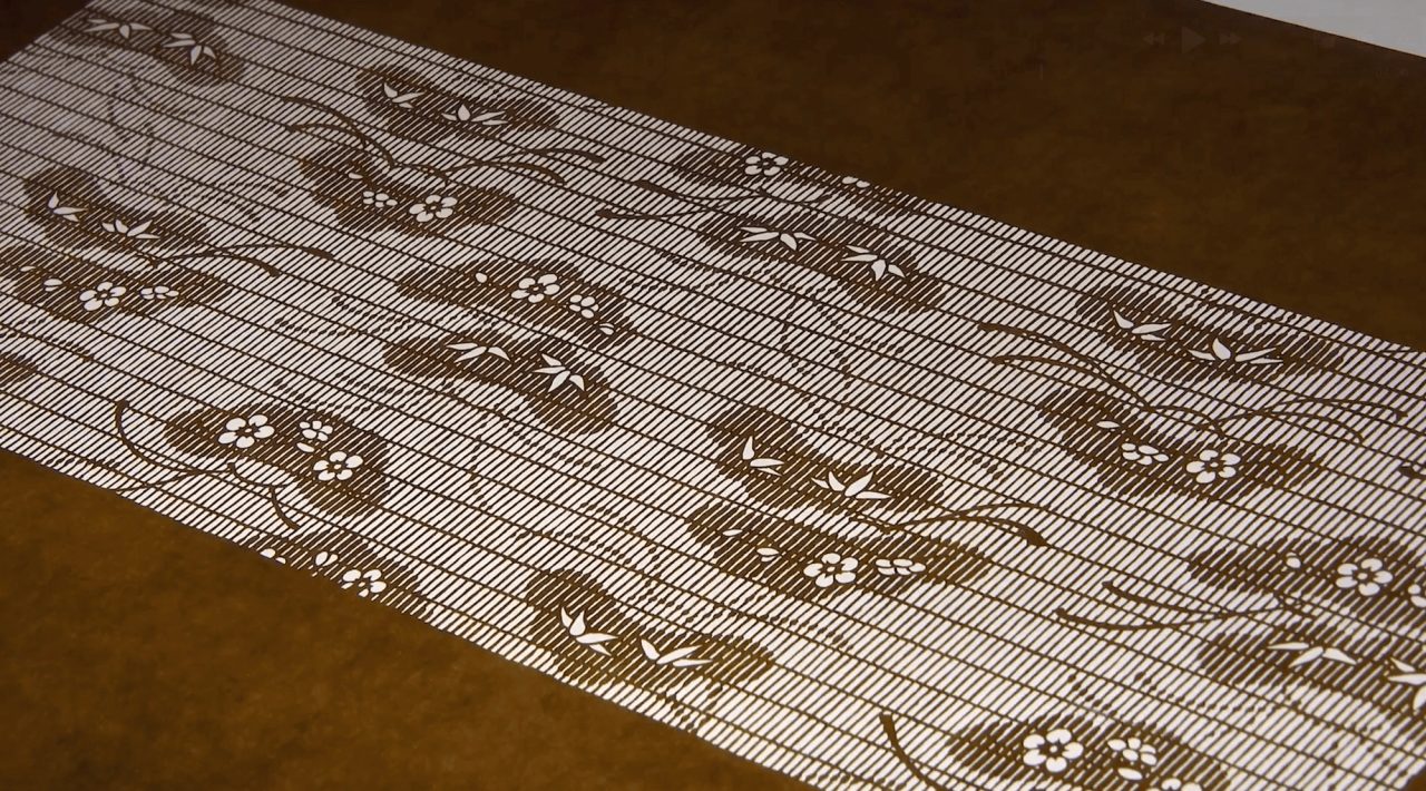 A long stencil of dark mulberry paper laid on a brown surface, cut through with a plum blossom and grass pattern over fine horizontal lines.