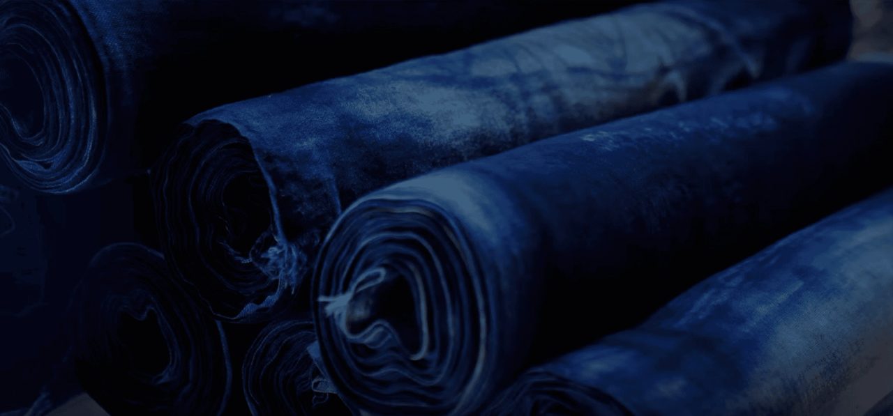 Bolts of cloth dyed deep indigo, rolled and stacked on their sides, the folds catching light in bands of lighter blue.