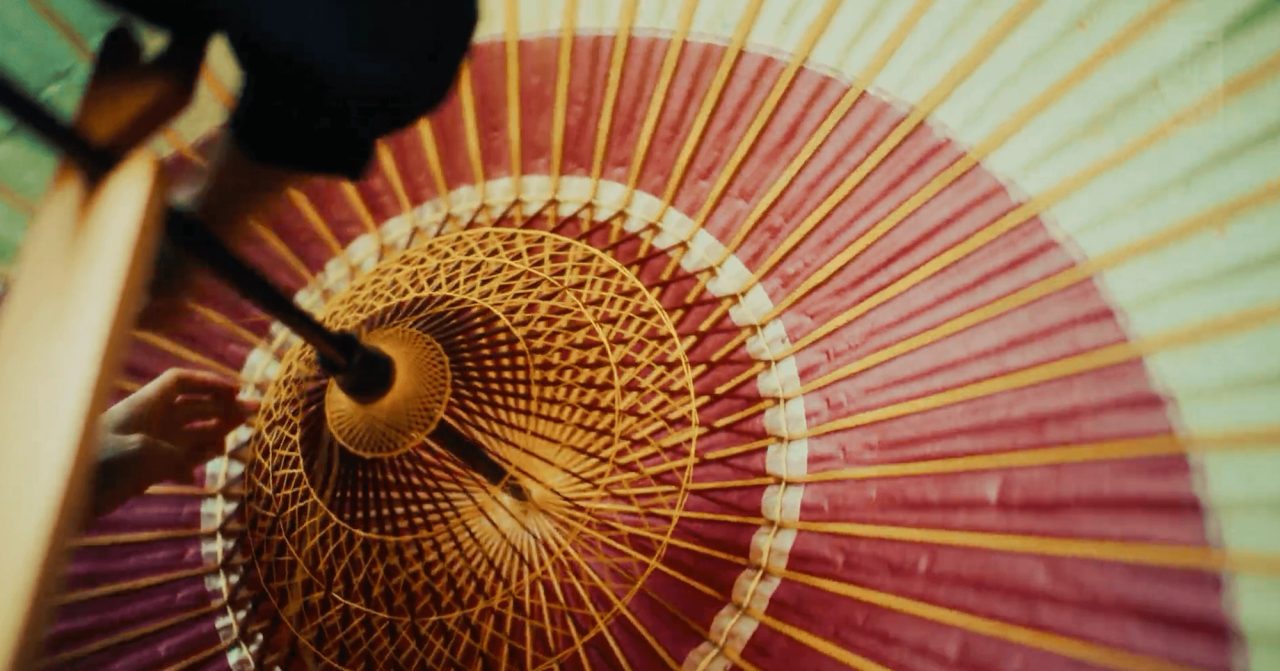 The underside of an open paper umbrella, bamboo ribs radiating from a threaded wooden core over deep red paper, with a hand adjusting one rib at the left.