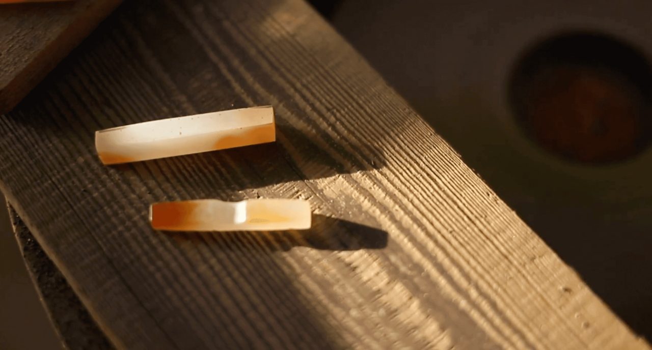 Two small polished agate blanks, one milky white and one banded orange, lying on a grooved wooden bench in low light.