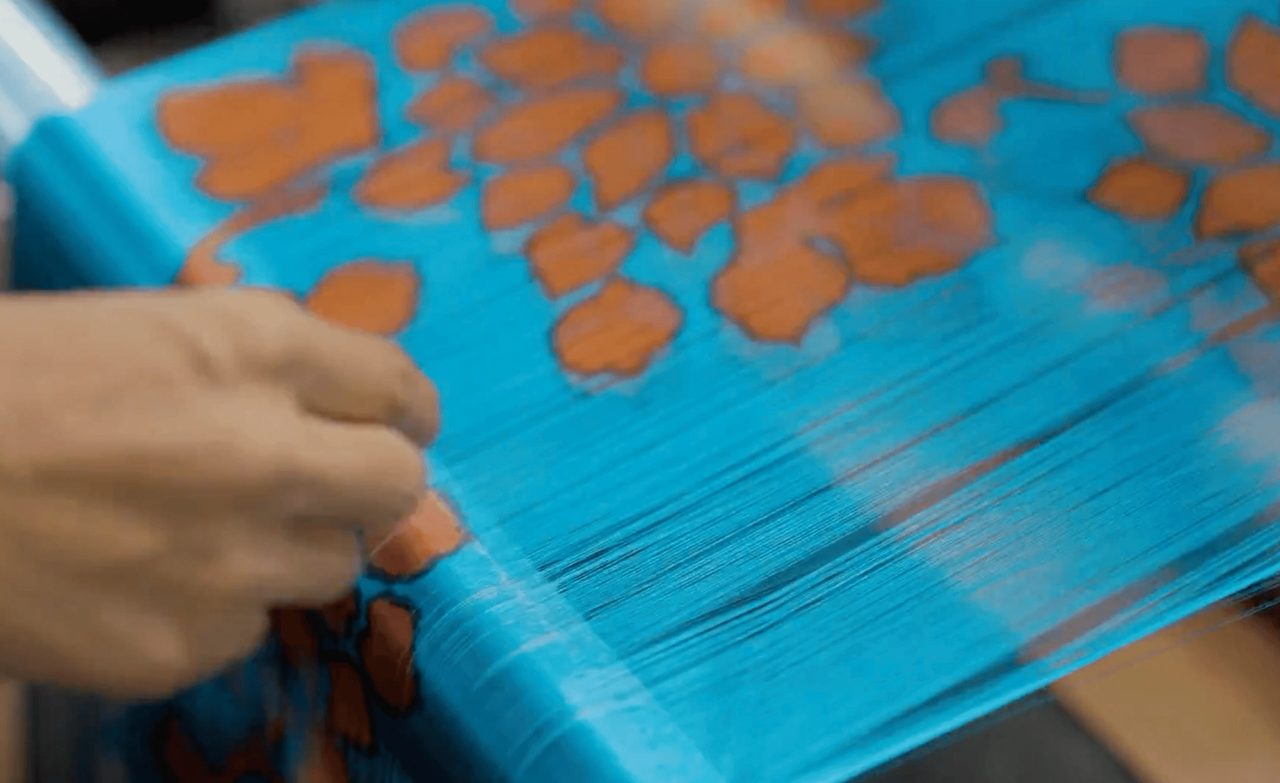 A hand smoothing a warp of bright blue threads stretched across a frame, printed along their length with orange leaf shapes.