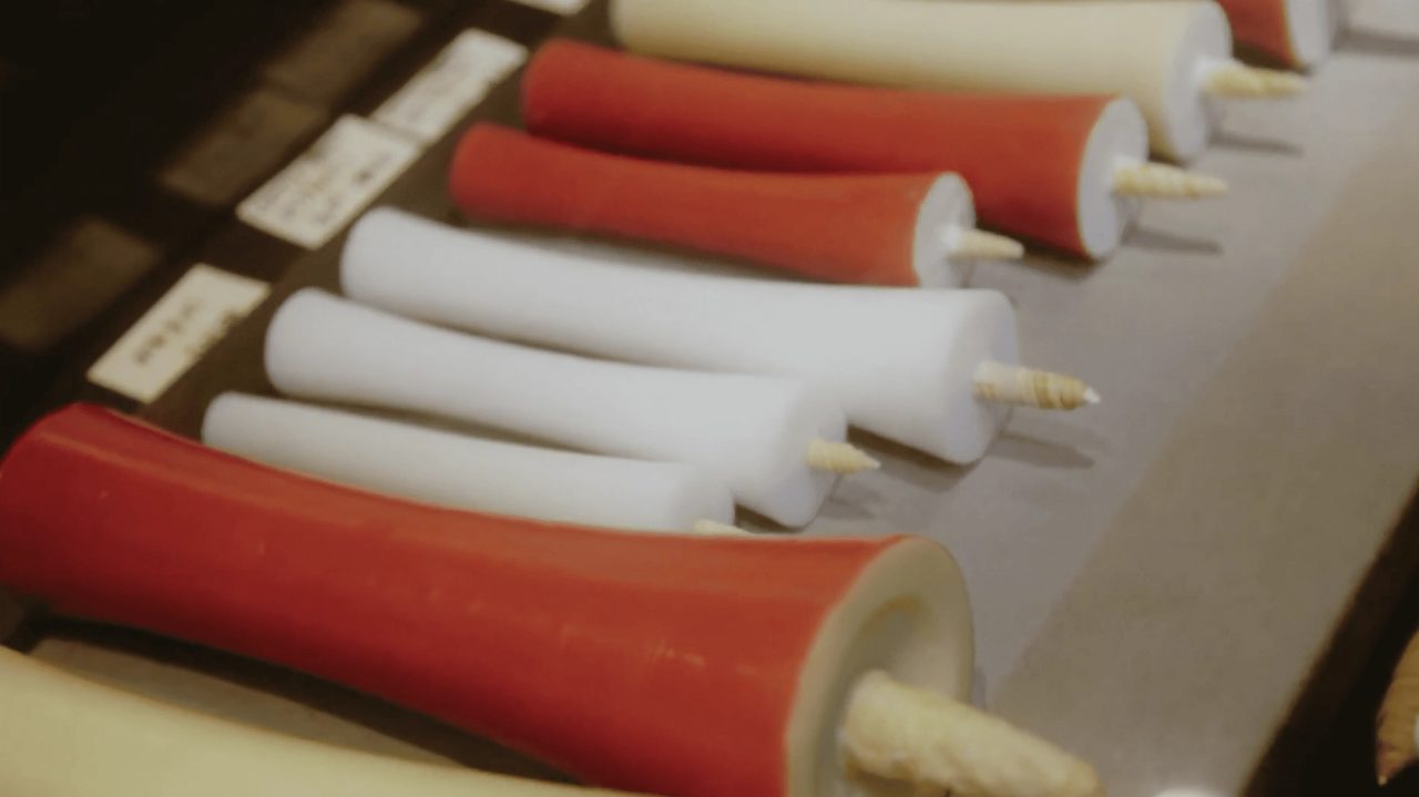 A row of hand-rolled candles lying on a bench, alternating deep red and white, each flaring outward at the top with a thick coiled wick.
