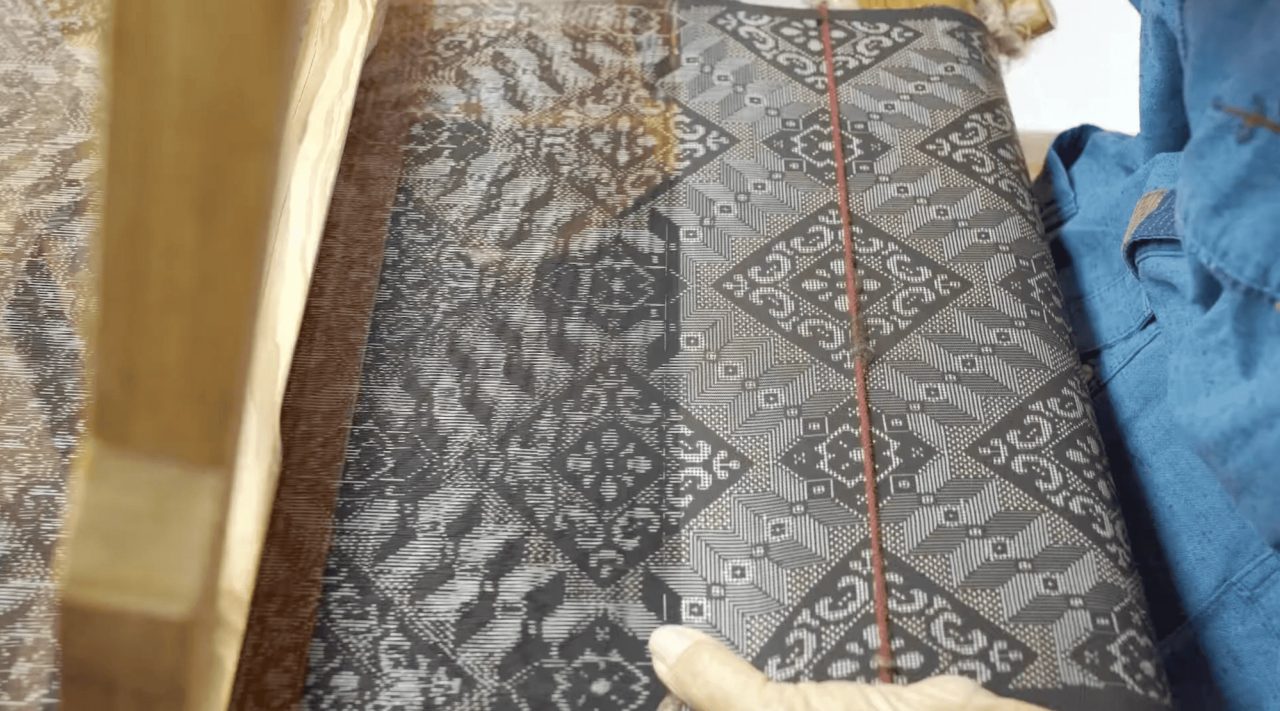 Cloth on a loom patterned in fine black and white diamonds and geometric motifs, with a hand resting at the lower edge and a red guide thread across it.