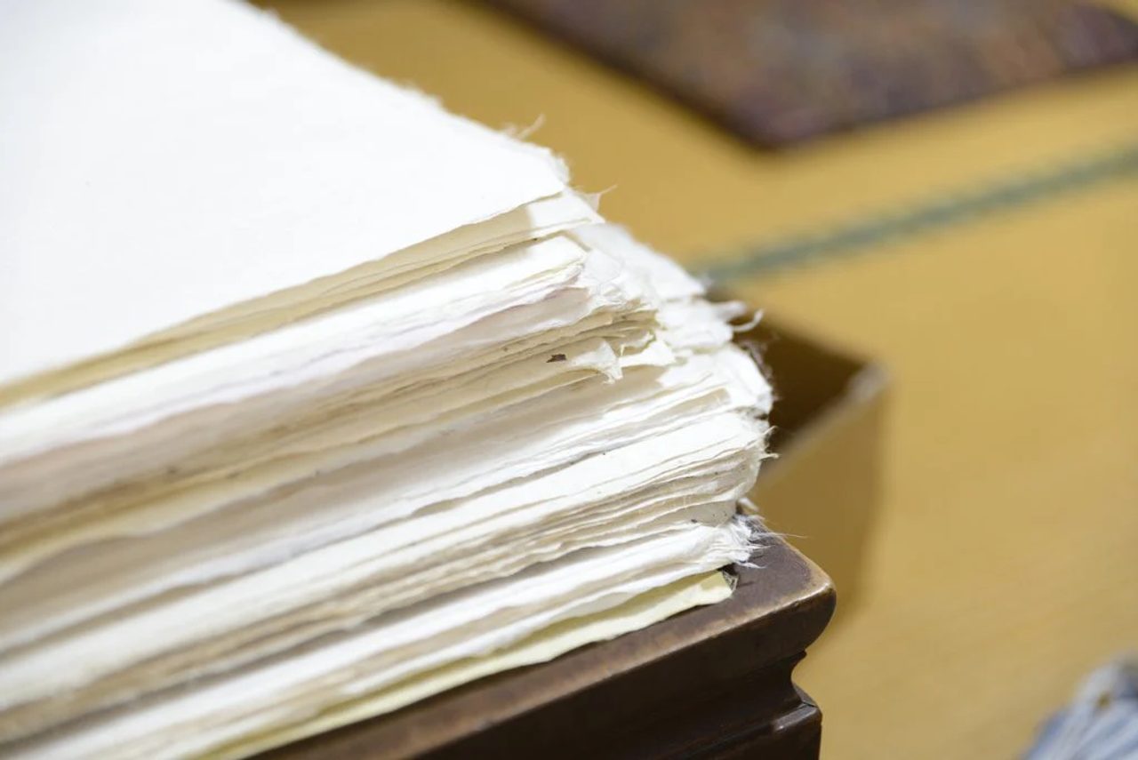 The torn edge of a thick stack of handmade paper sheets resting on a wooden board, the fibers visible along every deckle edge.