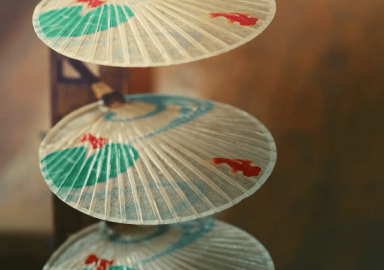 Two translucent round fans on slender handles, painted with green fans and a red goldfish, hanging one above the other against a warm background.
